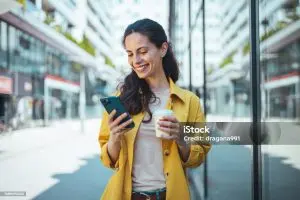 Smiling woman in a yellow coat looking at her phone and holding a coffee on a city street
