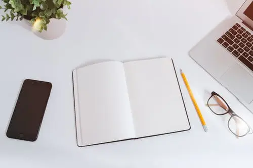 Open blank notebook with a pencil, glasses, phone and laptop on a white desk